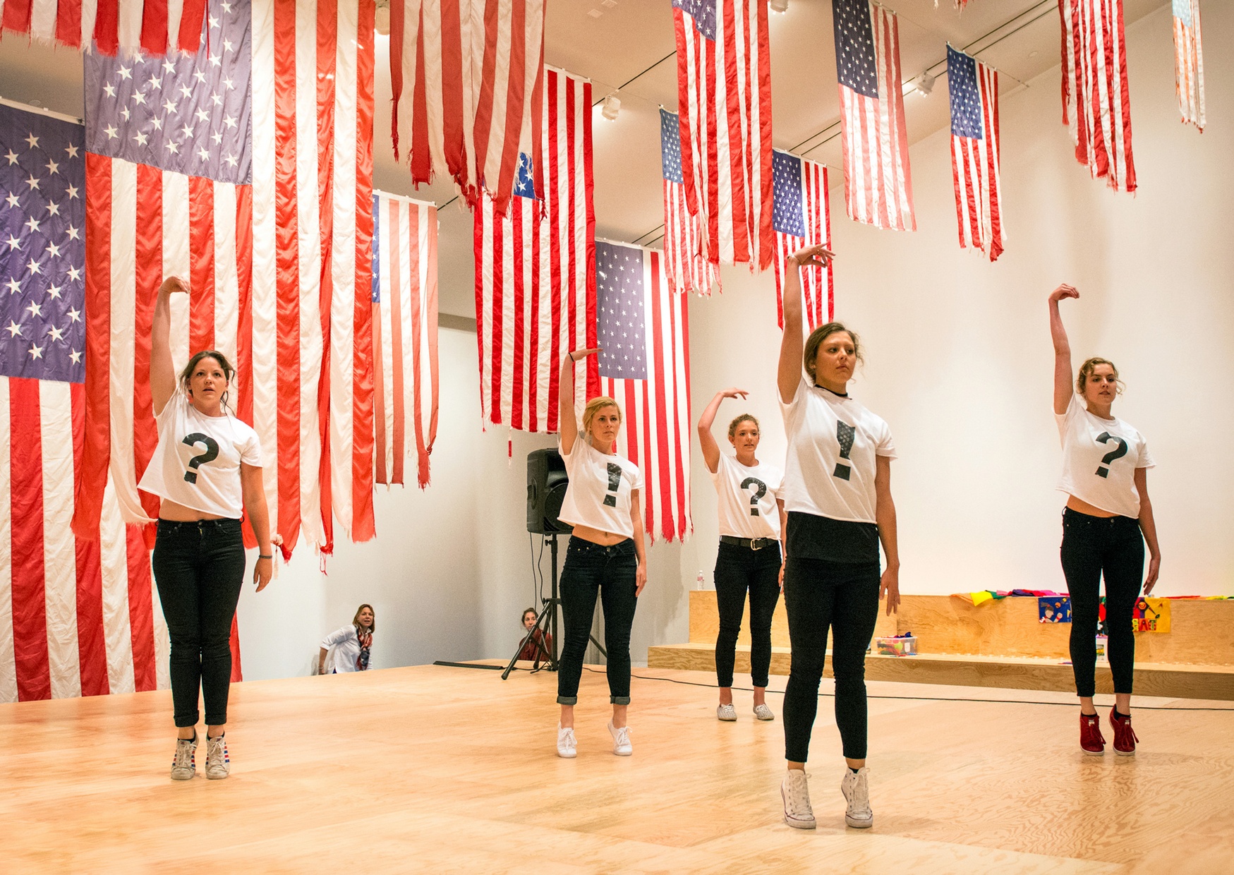 Five light-skinned females pose with one arm raised on a wooden stage surrounded by American flags hung from the ceiling.