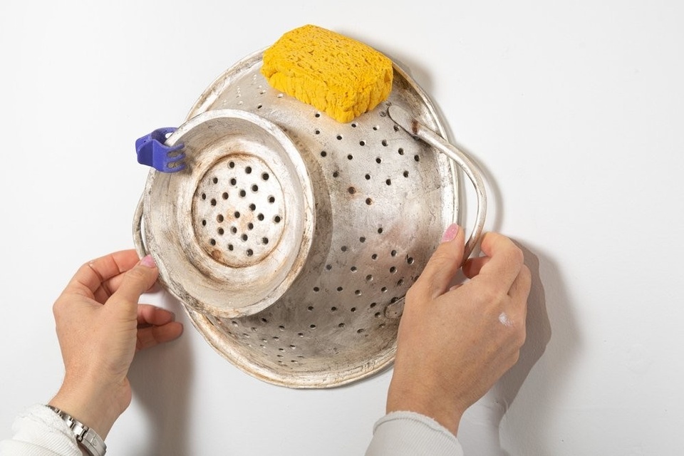 Two hands hold a silver colander against a white wall. The colander has a yellow dish sponge stuck to one side and a hair clip clipped to its base.