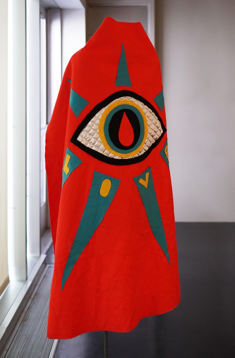 A long, red cape with a large eye, made of red, black, blue, yellow, and silver fabric; blue triangles, bearing yellow letters “L O V E,” radiate from the eye.