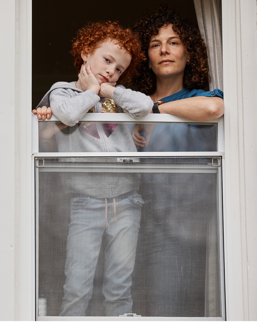 A photograph of a light-skinned woman and child look out a partially open window framed in white.