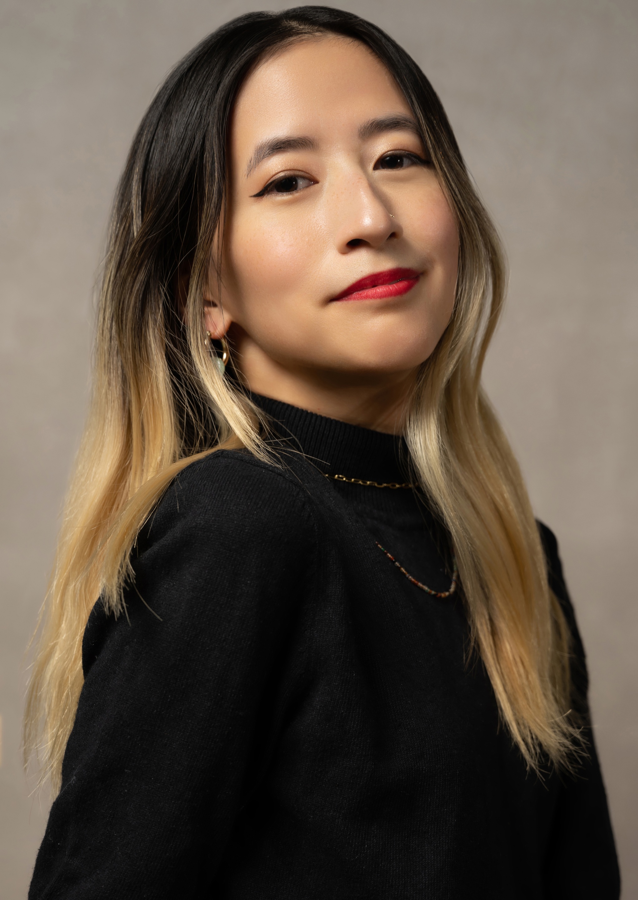 A portrait of Yo-Yo Lin: she is a Taiwanese American femme with long bleach blonde hair. She wears a red lip and smiles serenely at the camera in front of a tan studio backdrop. 