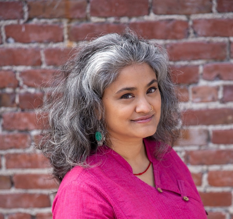 A photo of sujatha baliga standing against a red brick wall. baliga wears a bright pink shirt and has gray and black hair that falls behind her shoulders. 