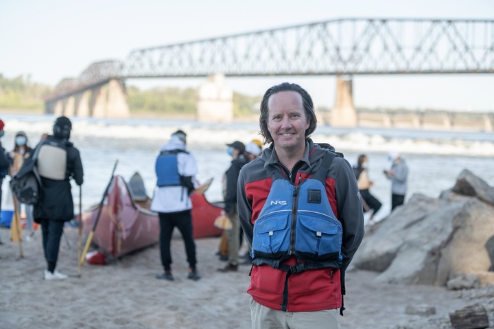 Person with blue life jacket stands in the foreground, in front of the waterfront and bridge. In the background, students prepare to get into canoes.