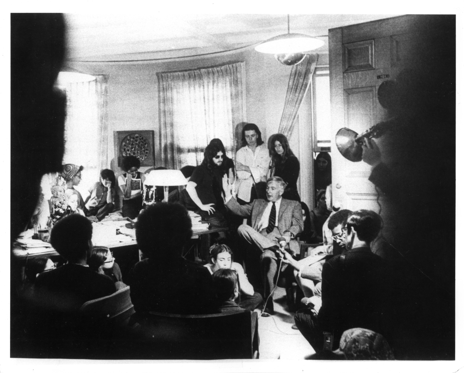 A black and white image of a group of light-skinned and dark-skinned young people in a room standing around an older light-skinned man seated in a chair with his right hand raised in oratory gesture.