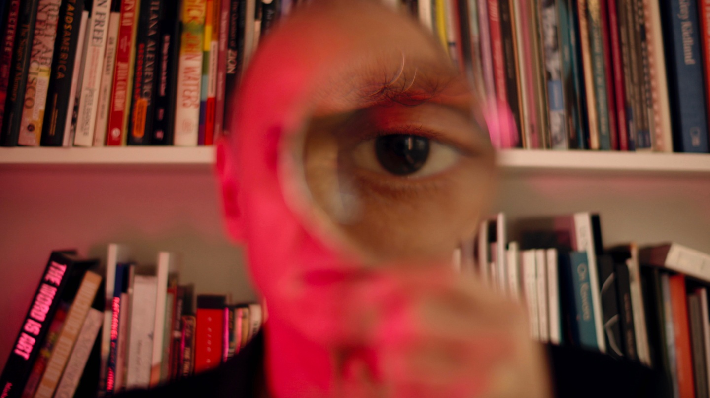 A person in front of a bookshelf holding a magnifying glass, their eye magnified. Red light from the left illuminates them.
