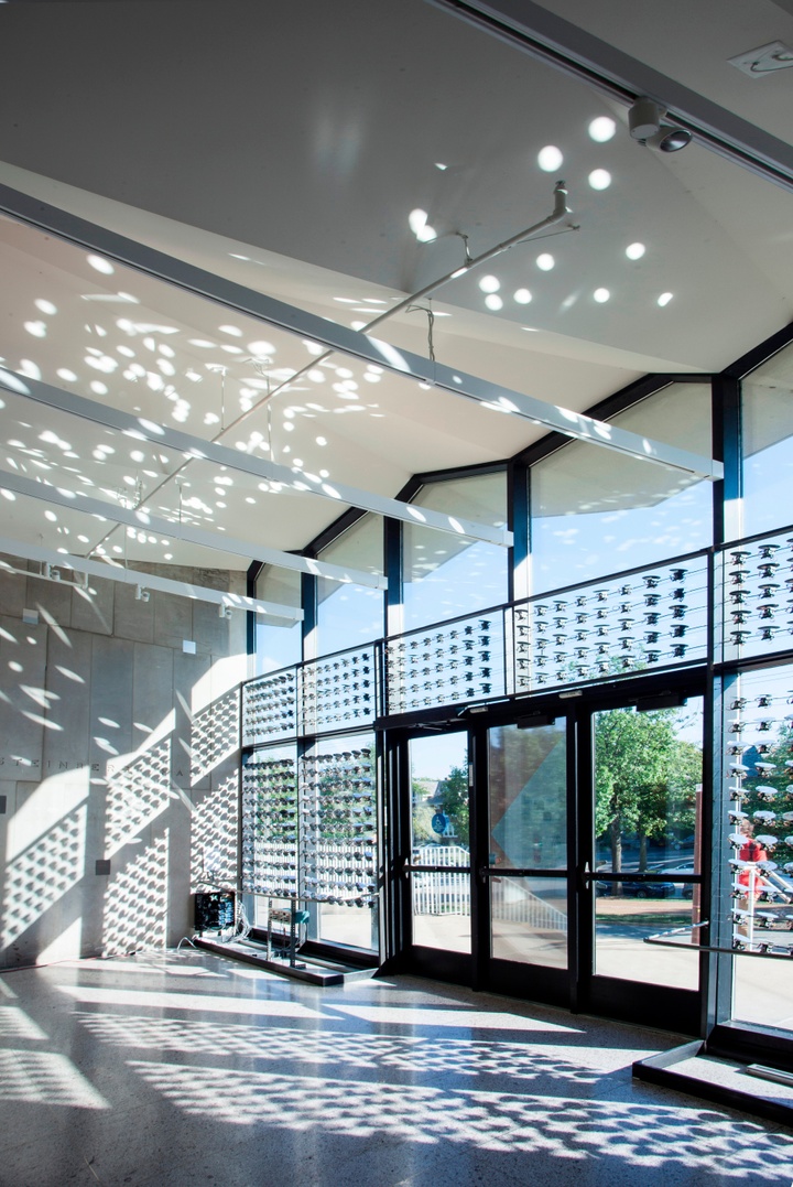 Open lobby area of Steinberg Hall, with reflections of light on the walls and ceilings, cast by a series of small mirrored surfaces hung along the floor-to-ceiling glass windows and doors on the back wall.