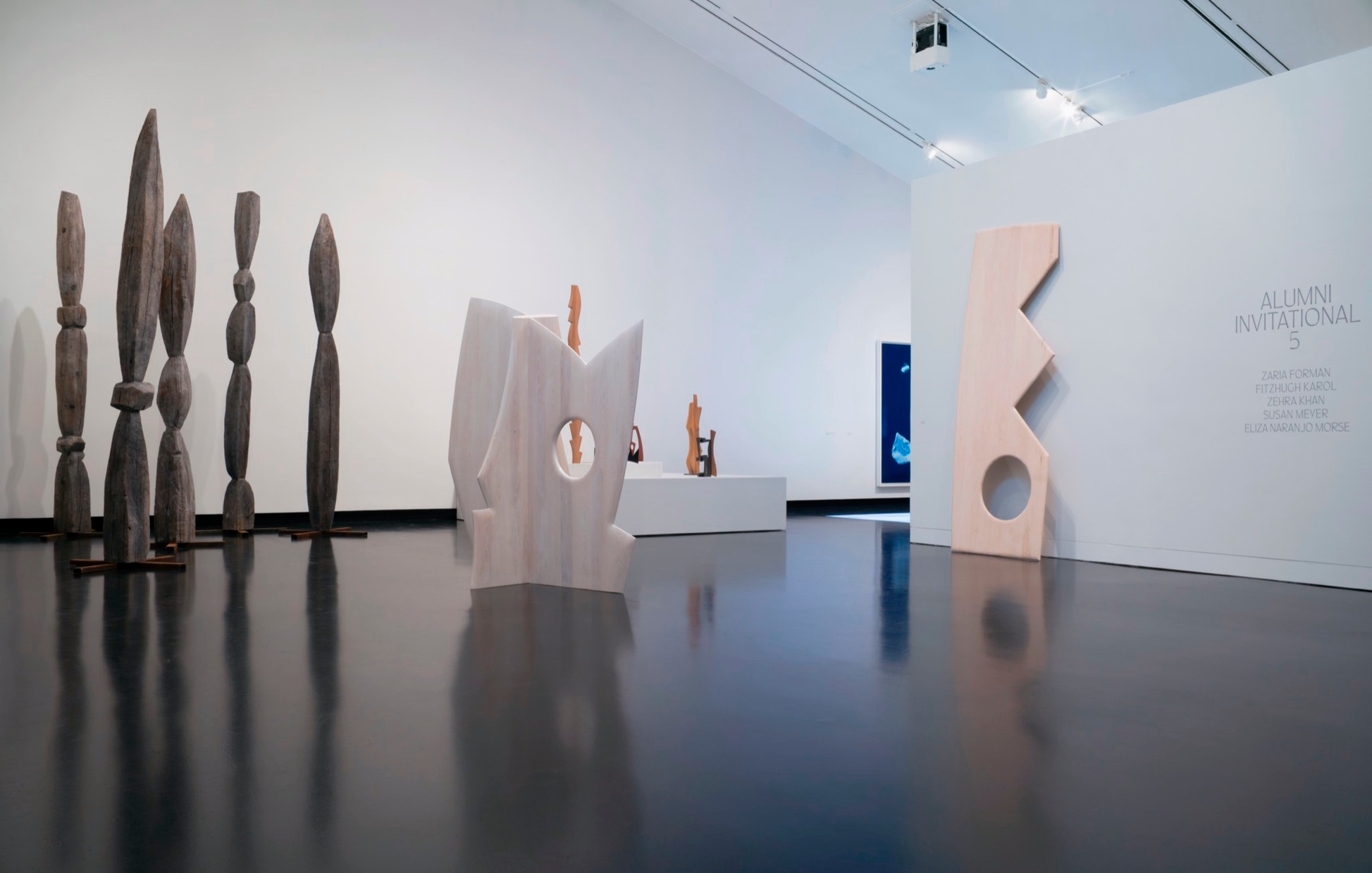 In a museum gallery with white walls and a black floor stand many geometric wooden sculptures.