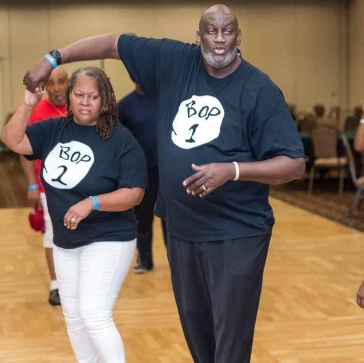 A photo of Audrey and June Donaldson, a Black couple who are dance instructors. They wear black t-shirts that say Bop 1 and Bop 2. Audrey spins June on a wood dance floor.