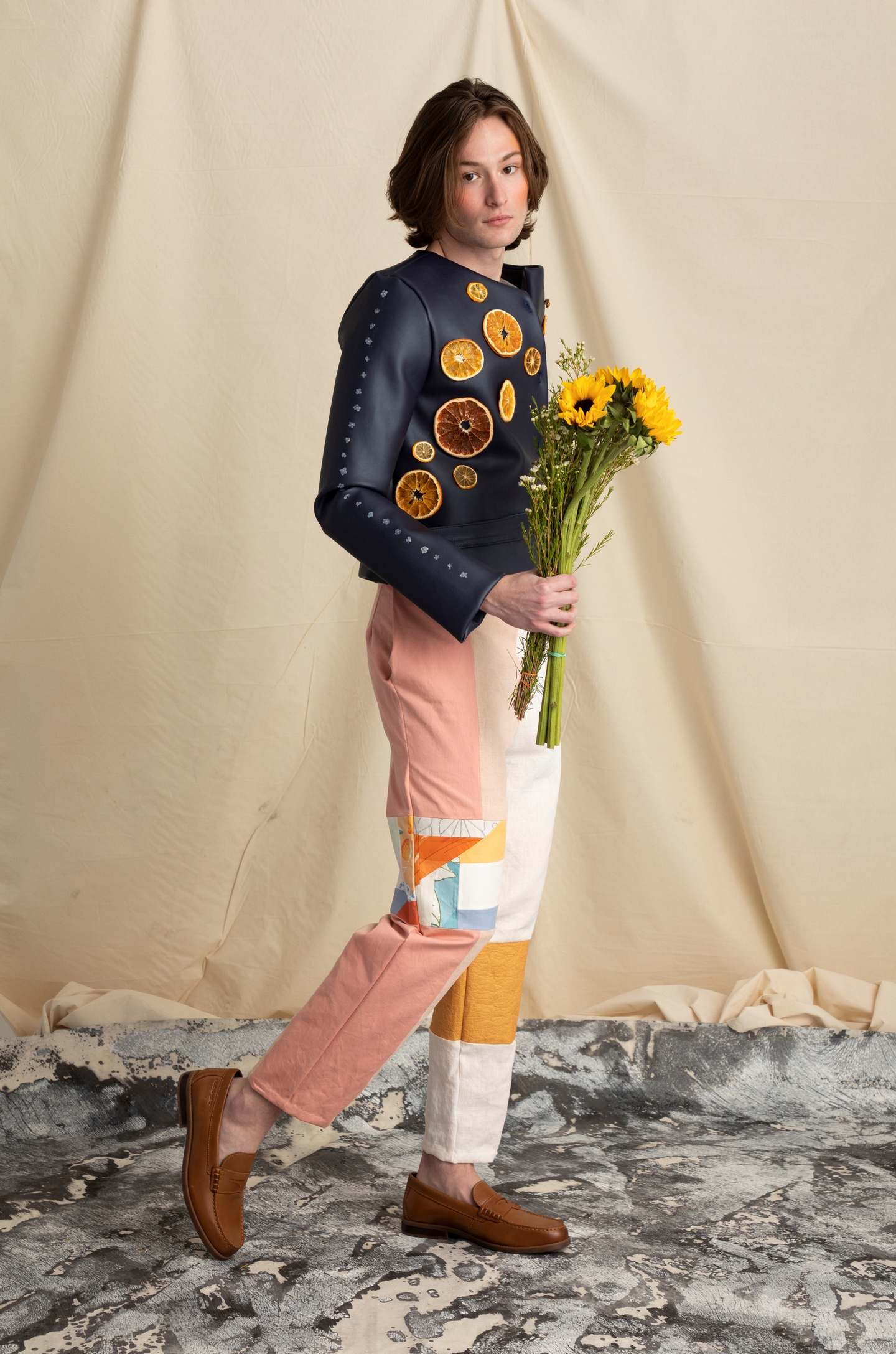 Model wears a black fake leather jacket studded with dried orange slices and narrow leg pants made of quilted pieces of pale pink, white, beige, and yellow fabrics. The model wears brown leather loafers and carries a bouquet of yellow flowers.