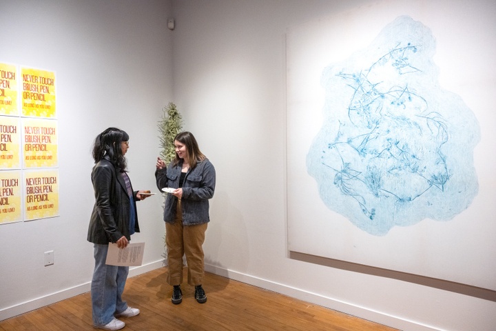 Two students chat in the corner between two artworks. To the left are a set of yellow letterpress prints that read "Never touch brush, pen, or pencil as long as you live!" To the right is a pale cyan-tinted print of foliage arranged in an amorphous shape.