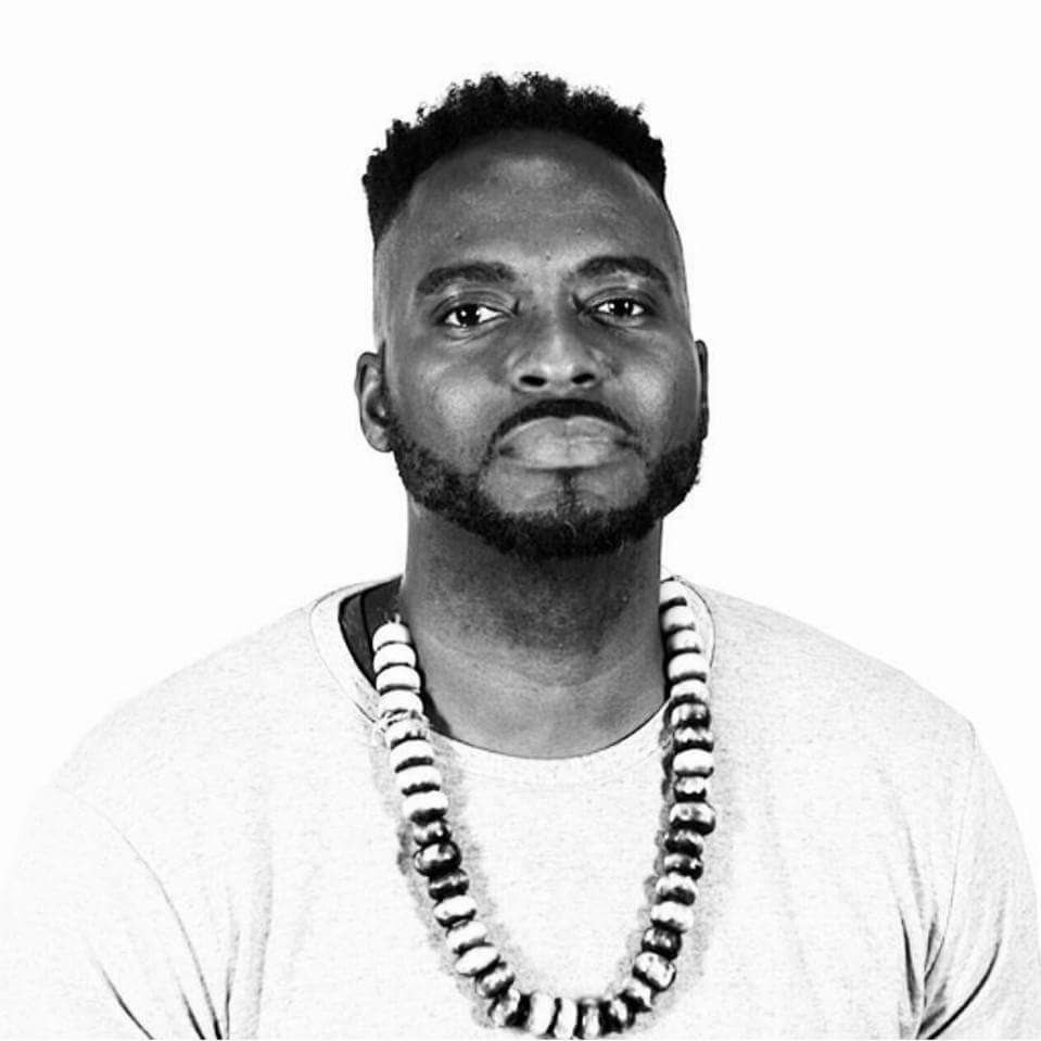 A black-and-white photo of Jason Minnis a.k.a. ClassicBeatz sitting against a white background. Minnis has a mustache and beard low along his jaw and wears a necklace of large disc-like beads. 