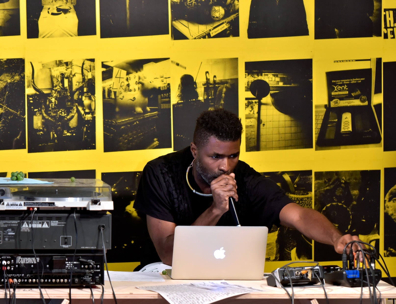 A dark-skinned man holding a mic close to his mouth looks at a laptop and adjusts a soundboard. He sits in front of a yellow wall with different scenes screenprinted in black.