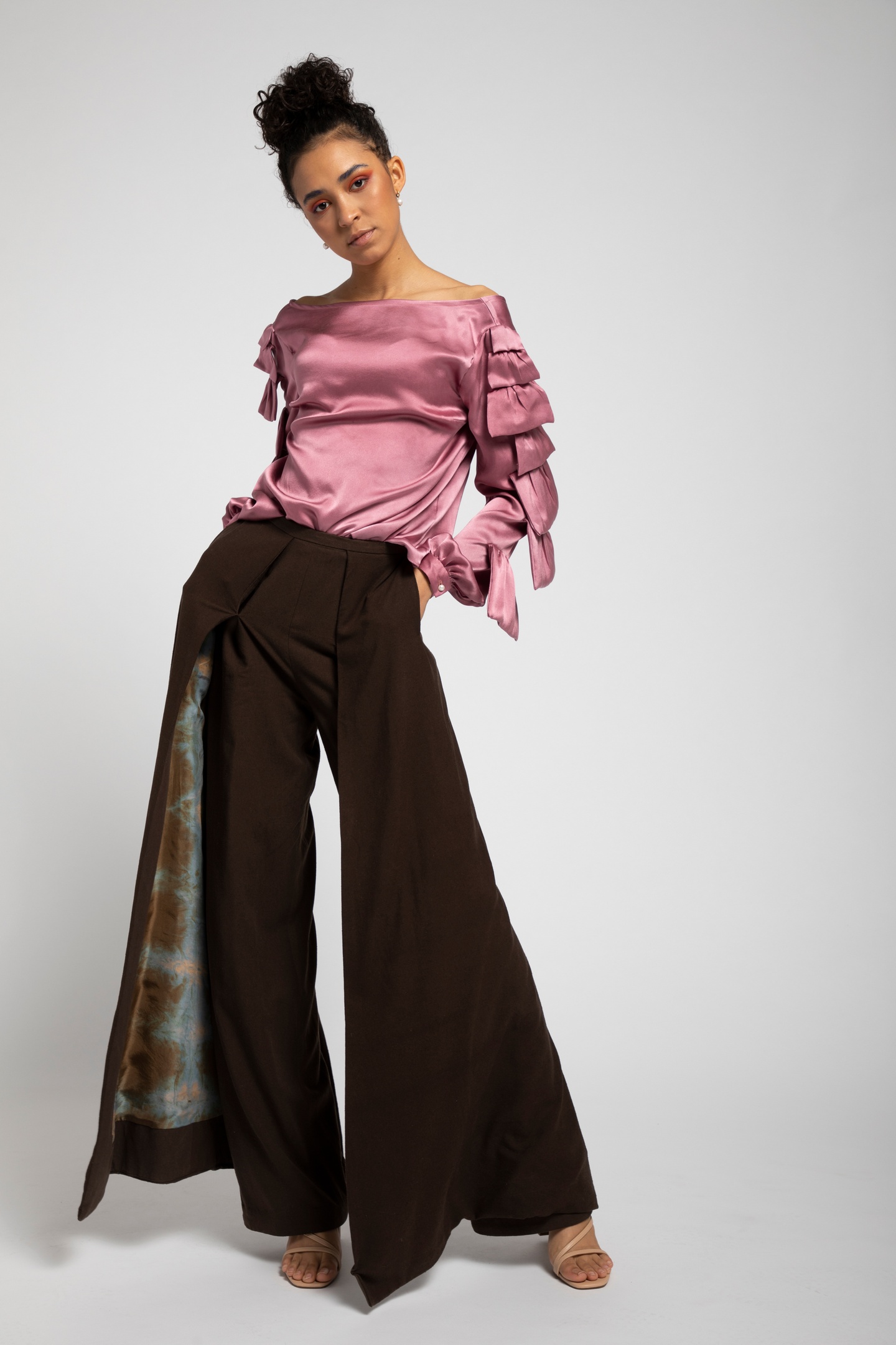 Model in a mauve satin blouse with a wide collar and ruffled long sleeves, paired with dark brown trousers with outer skirted panels lined with a blue and gold patterned satin.