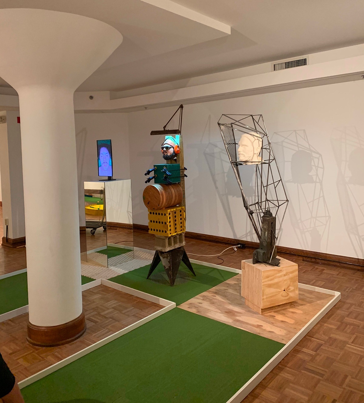 Miniature golf hole installed in an indoor gallery space. The hole design features three tall sculptural elements, two featuring scultped heads and the third featuring a digital version of a head.