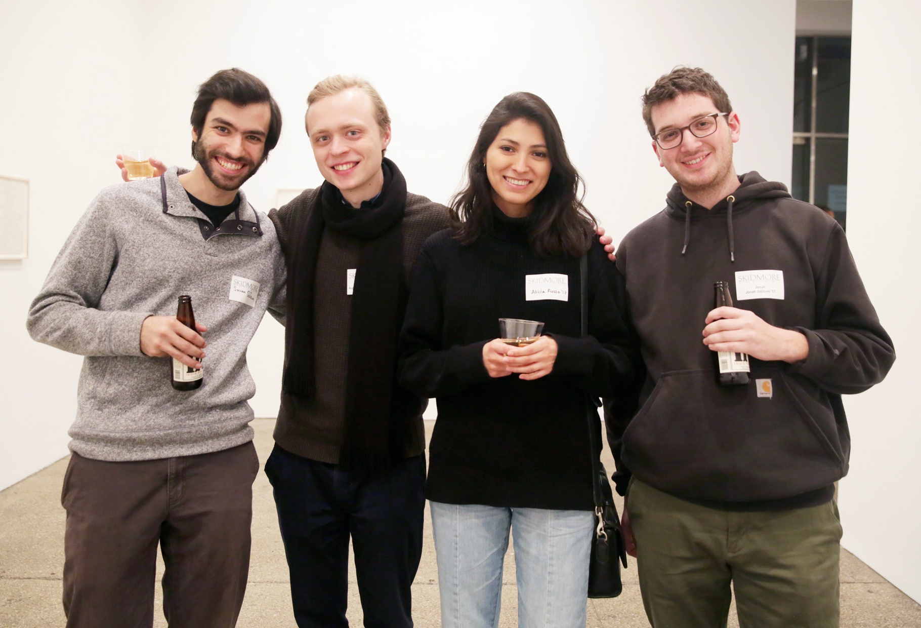 Four, young, light-skinned people stand next to each other smiling, holding drinks in front of a white wall.