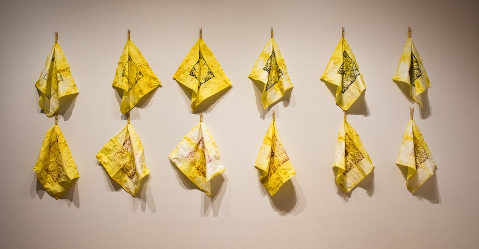 Two neat rows of six mottled yellow kerchiefs with black ink designs are hung with clothespins by their corners on a wall.