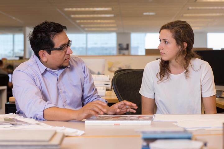 Two people sit at a table in an open office and discuss something.