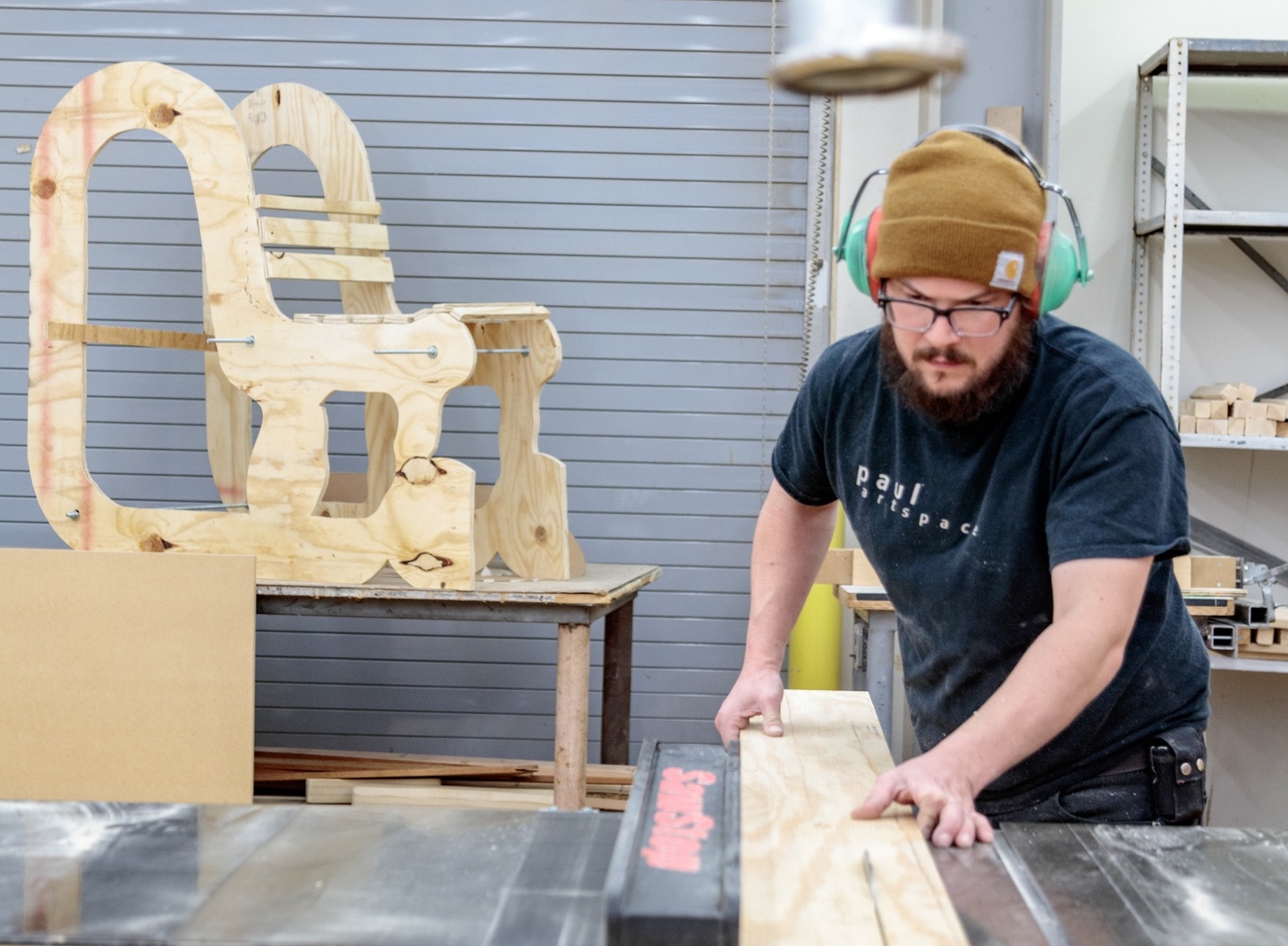 Person in a beanie and earphones uses a table saw in a shop space.