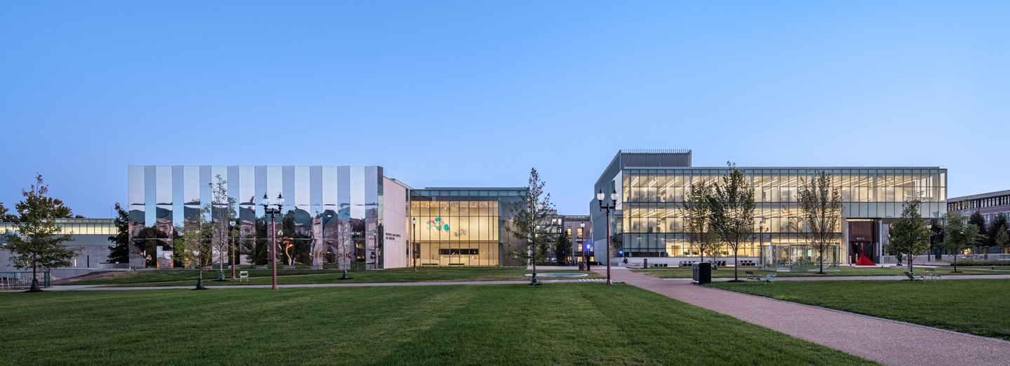 Wide angle shot of two buildings on a college campus. The left one has a pleated exterior mirrored facade and the right is a 3-story glass walled building lit from within.