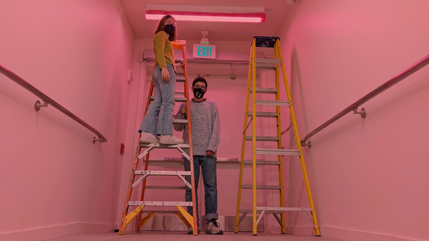 Two students wearing COVID-19 masks, standing by a pair of ladders in a hallway that's lit with a pink overhead light.