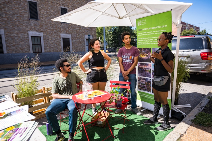 Four students chat underneath an umbrella set up in a street parking spot turned into a parklet.