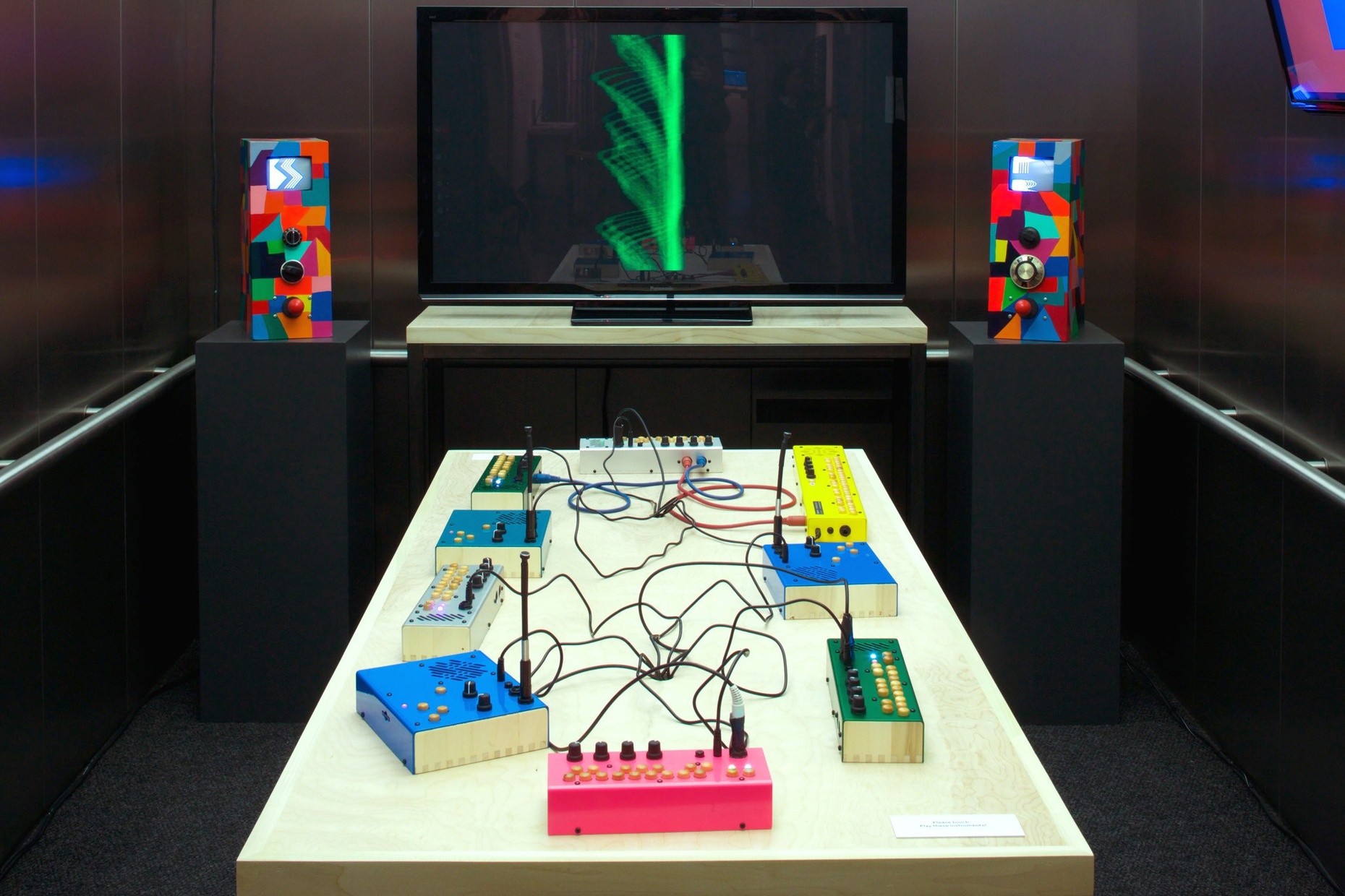 Colorful sound boxes connected by circuit wires rest on a white table in front of a TV and two abstractly colored speakers.