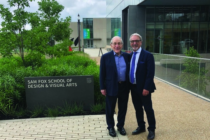 Two men dressed in dark suits pose for a picture on an academic campus in front of a dark gray granite sign that reads am Fox School of Design & Visual Arts. Behind them is lush green foliage and contemporary academic buildings made of metal, glass, and limestone.