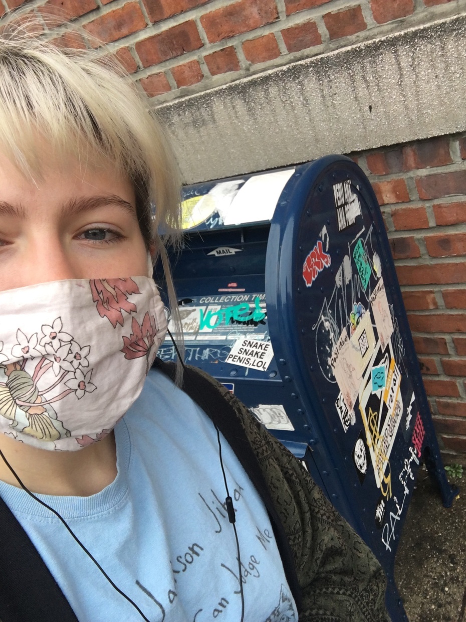 A blonde, light-skinned woman wearing a mask stands in front of a blue mailbox bin covered in graffiti and stickers.