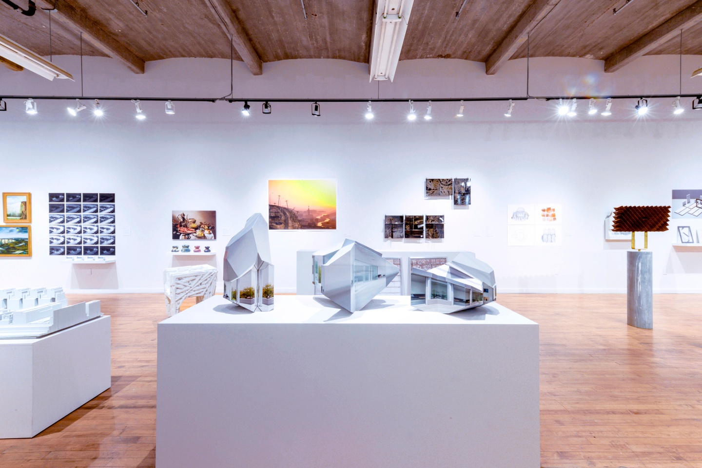 Installation photo of an architectural exhibition in a gallery space, with 3D models on white pedestals and 2D works on the walls.