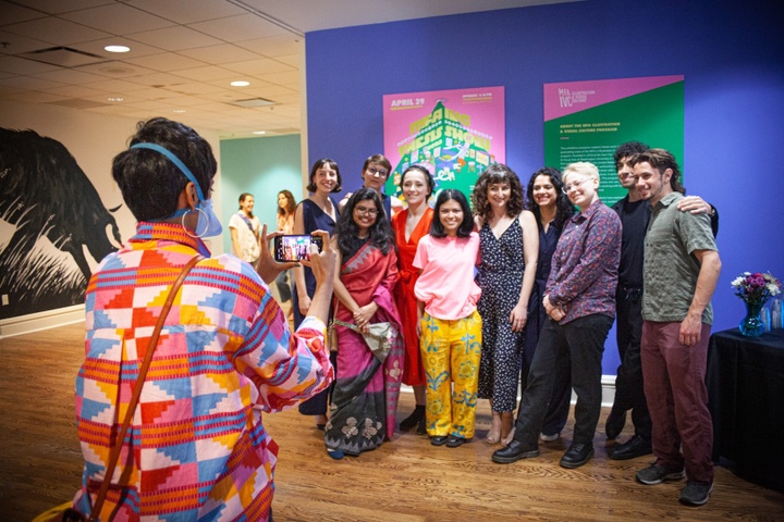 Group of smiling students poses in front of a blue wall hung with a bright pink and green MFA-IVC exhibition poster.