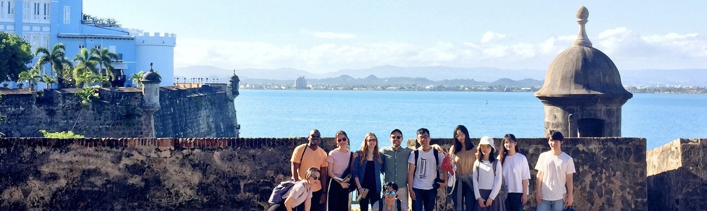 Group of students standing outdoors, in front of an old stone wall in San Juan, Puerto Rico, with the waterfront in the background.