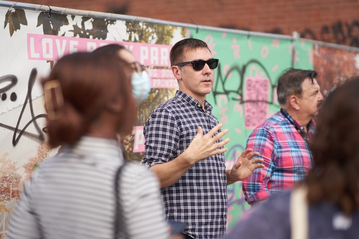 John Kamp and James Rojas stand in front of a construction fence at Love Bank Park and speak to a group of students.