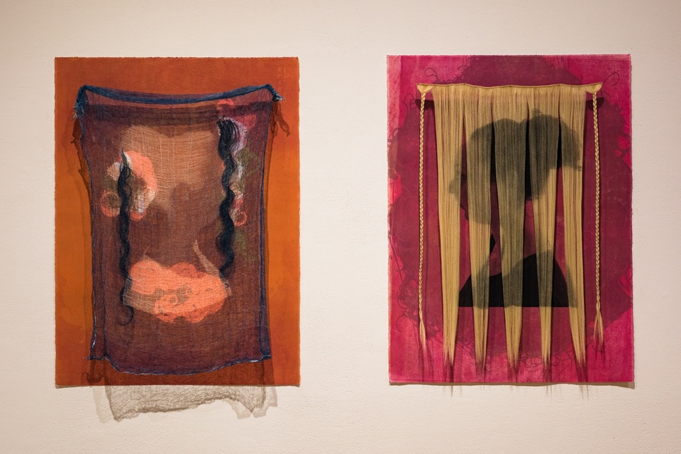 On the left is a vertical rusty orange panel with a blue cheesecloth and two long, dark, curly strands of hair tacked to it. Curly hair-like designs can be seen beneath the cheesecloth. On the right is a magenta vertical panel with a curtain of blonde hair screening the silhouette of a person's bust with a large hair bun.