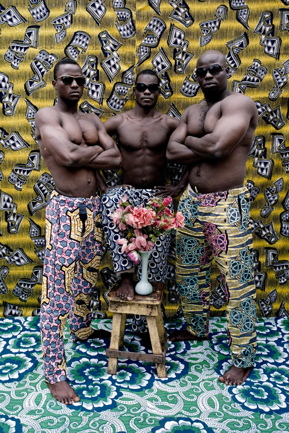 Three black men in sunglasses pose around a vase holding pink flowers. They are in front of a gold, black, and white backdrop and are standing on a floral fabric.