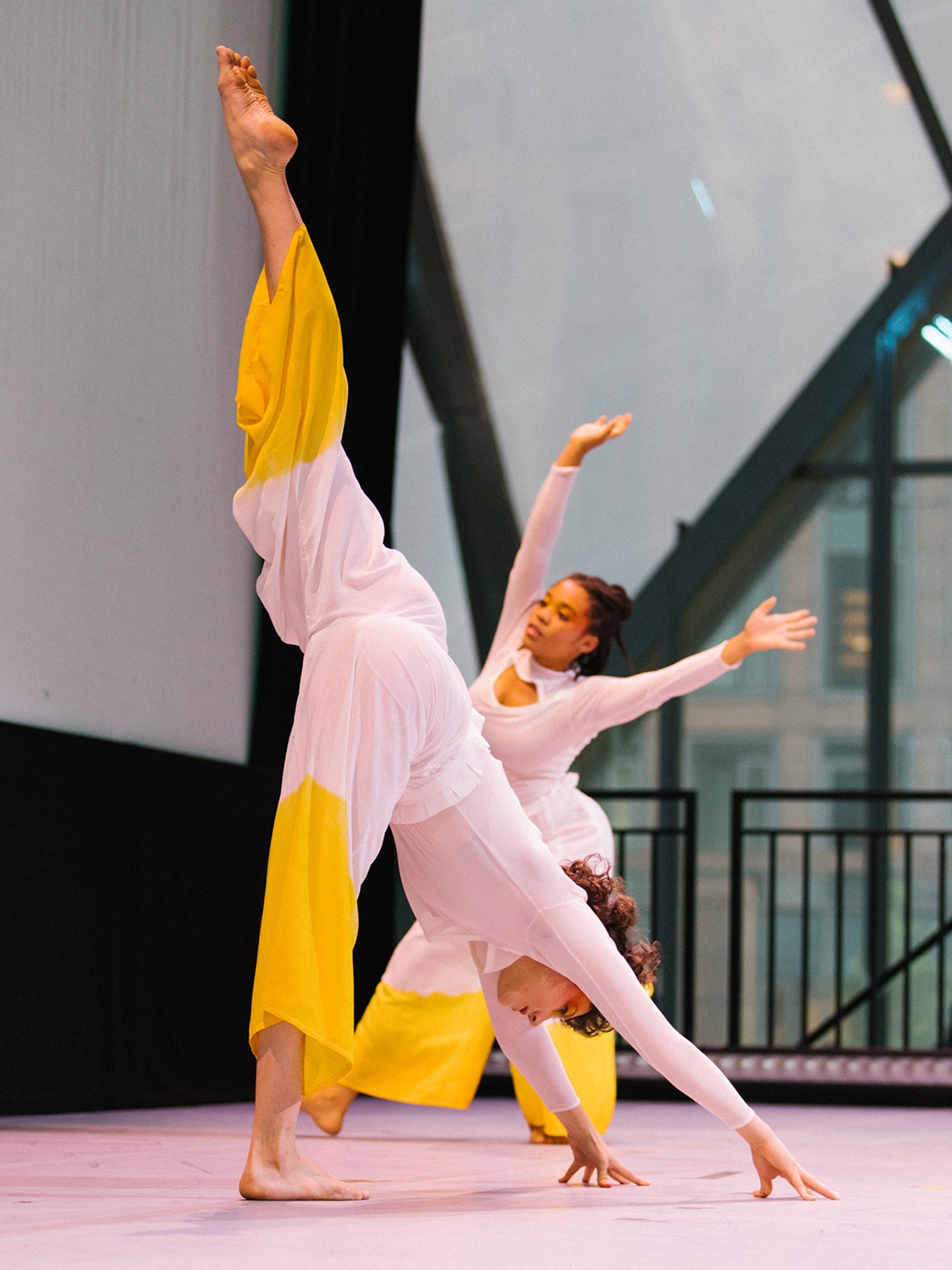 Two dancers on stage in white costumes with the pants dyed bright yellow. In the foreground one dancer bends over to touch the ground, lifting their left foot high into the air. In the background the second dancer extends both arms above their head.