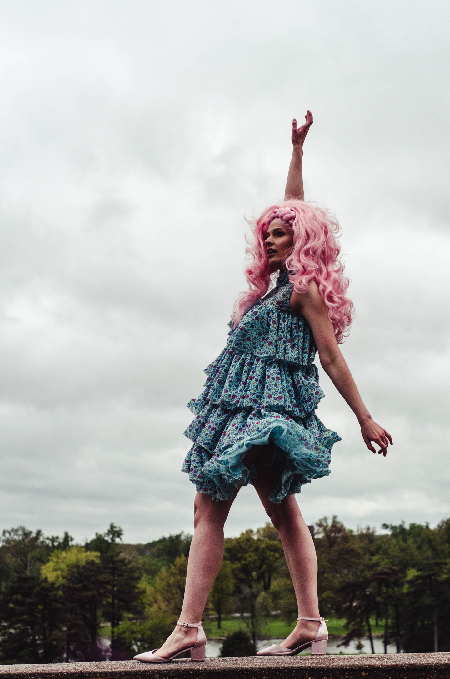 Model in a curly pink wig wears a kneelength sleeveless dress with four tiers of ruffles, made of a pale blue fabric printed with a colorful repeating planet icon pattern.