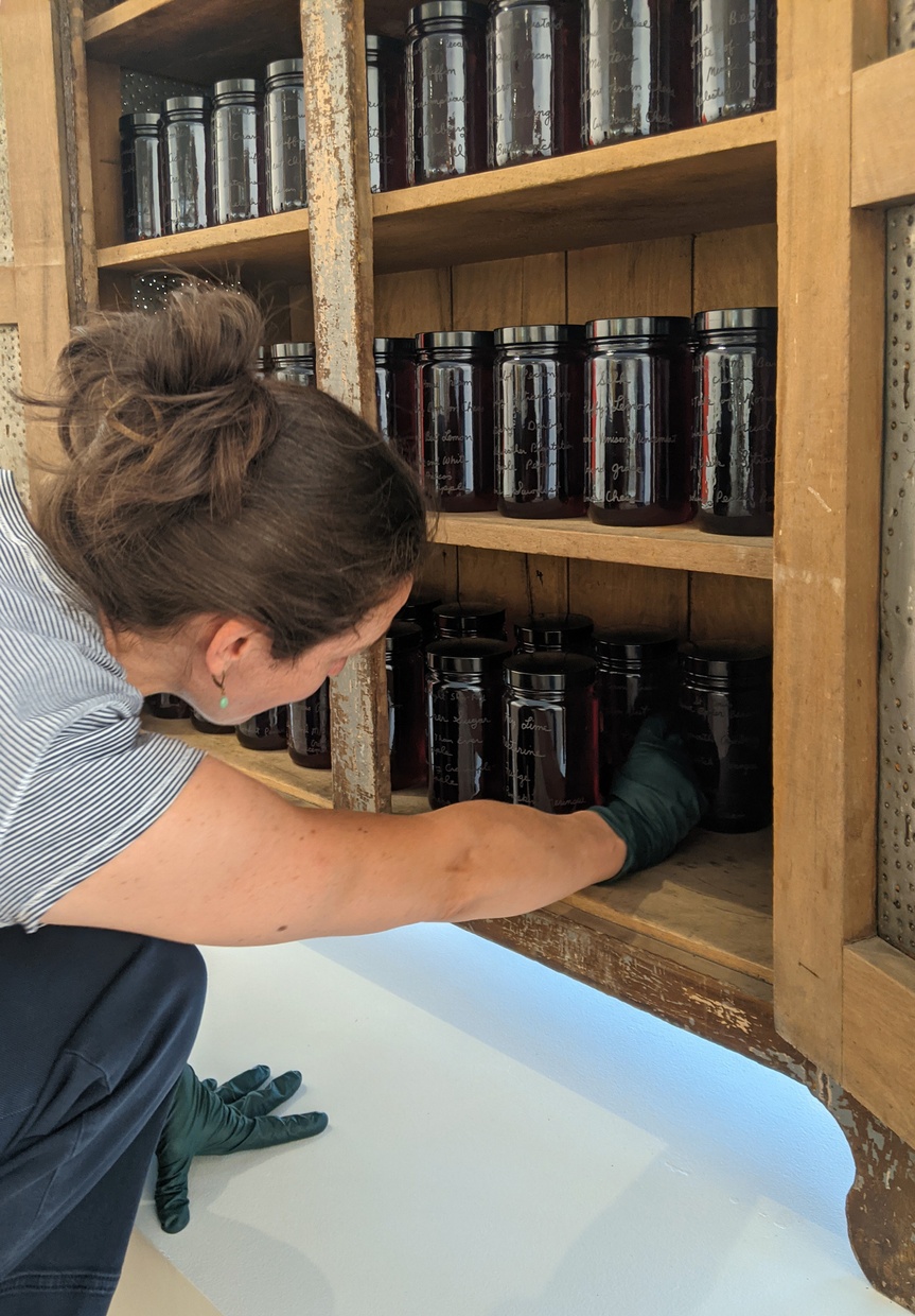 A woman is crouching and reaching into a wooden armoire of brown engraved glass jars.