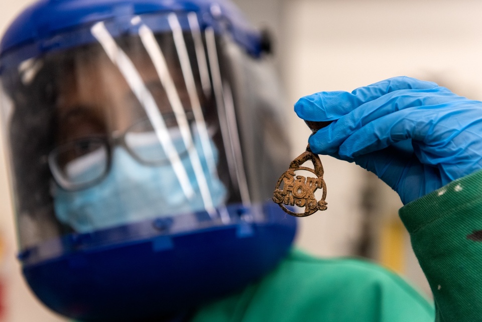 Student in a face shield and protective coat holds up a little bronze pendant with block letters reading "Sam Fox Shops."