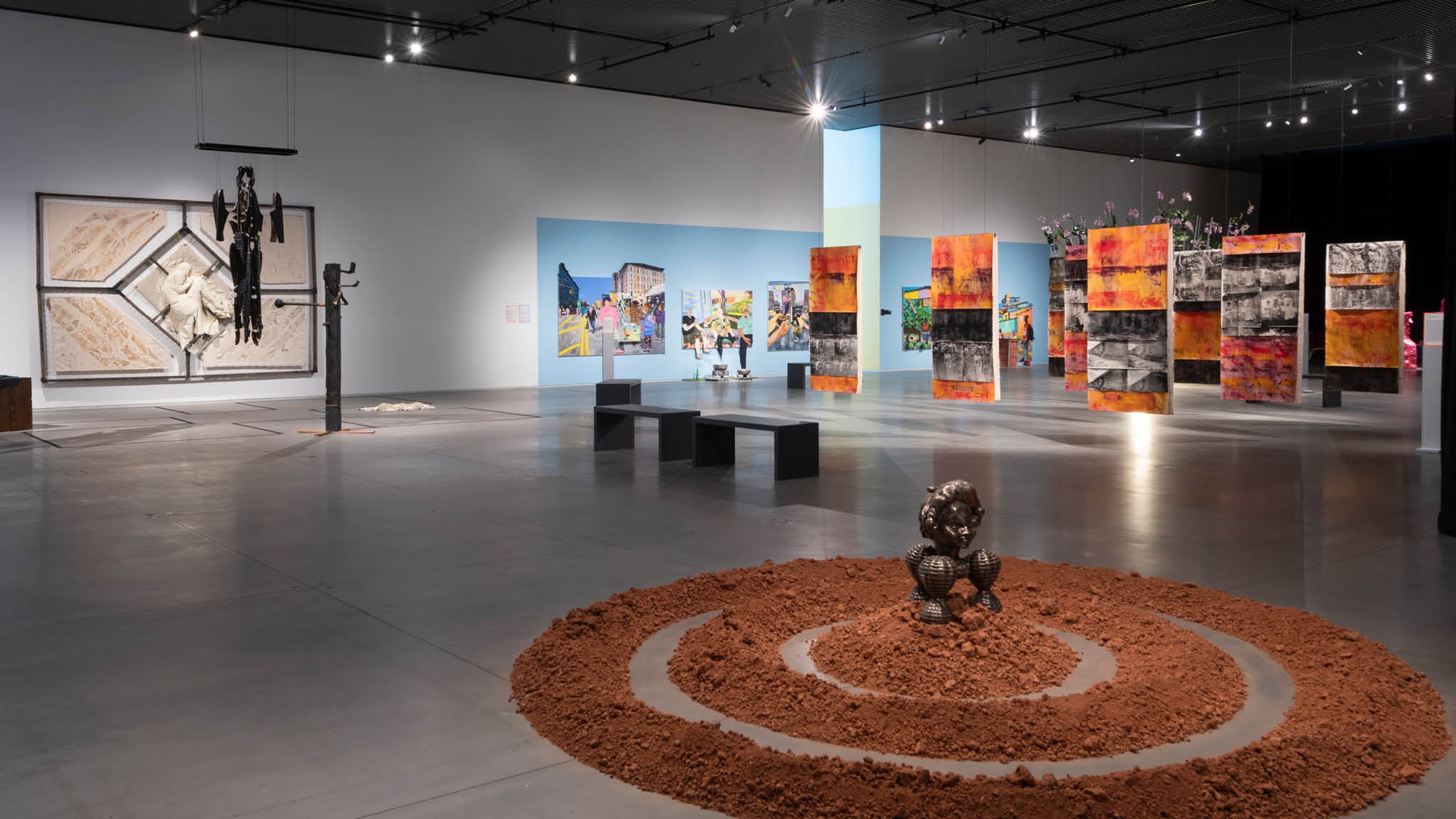 A view of an exhibition in a large art gallery without any walls. In the foreground is an artwork on the floor that consists of three concentric rings of red dirt. At the center sits a bronze sculpture inspired by the Benin Bronzes, historical artworks from Benin, present-day Nigeria. In the background are other paintings and installations. In the background are various paintings hanging from the ceiling and walls.