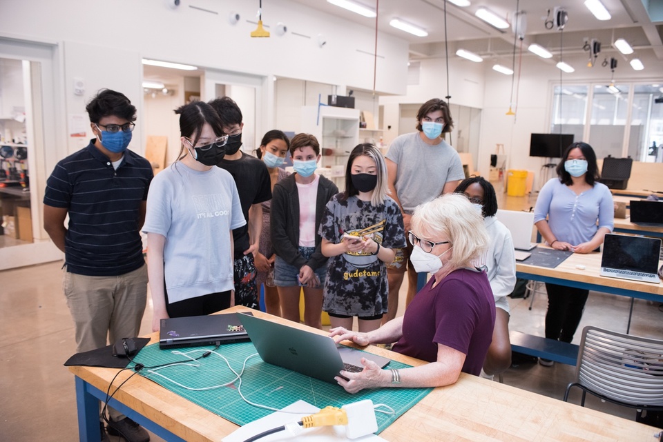 Group of students in a maker space gathers around an instructor demonstrating something on their laptop.