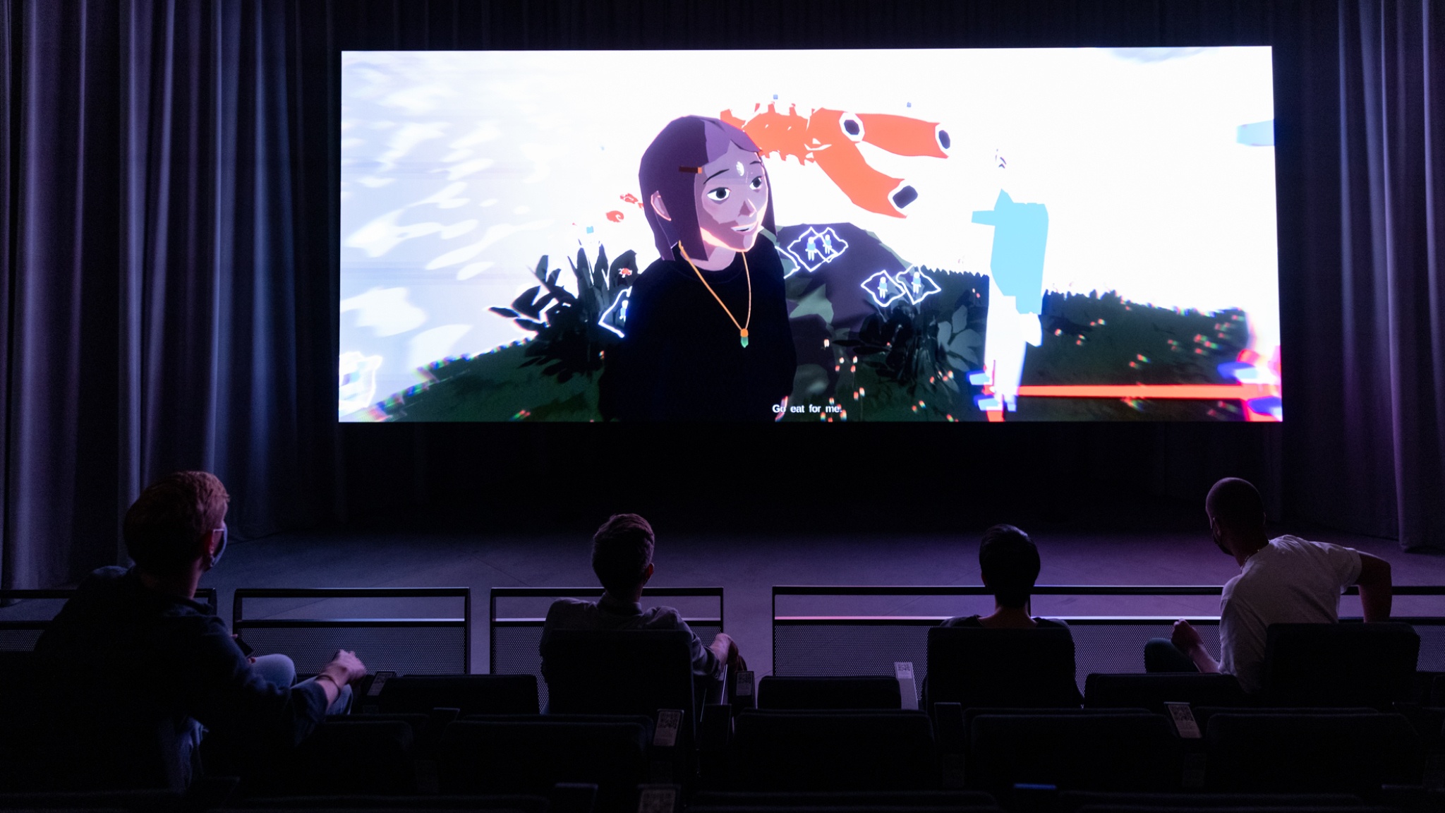A film screen with a scene from an anime projected on it, with in the foreground the back of four viewers' heads pictured from higher up in the cinema-style seating