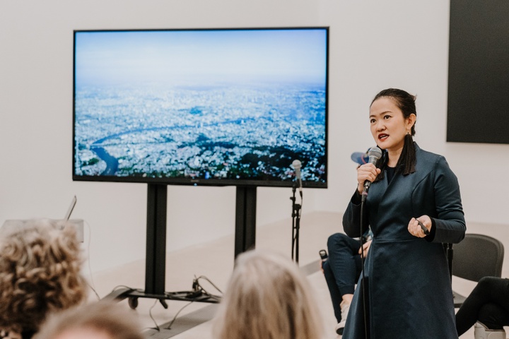 Voraakhom speaks in front of screen with misty aerial view of a river city.