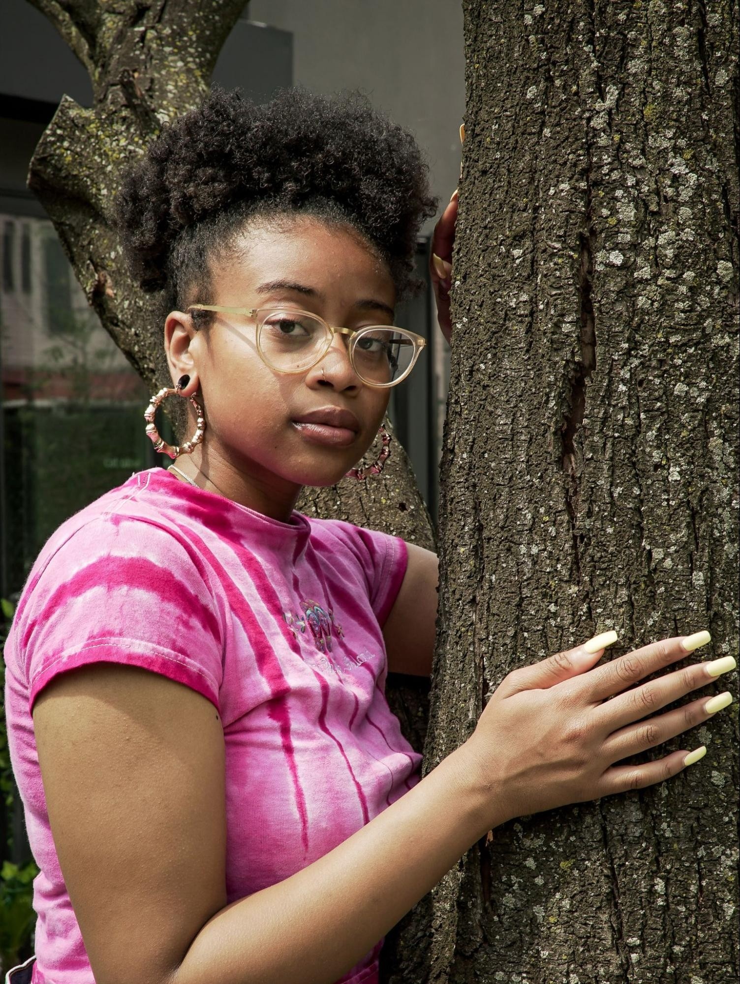 A Black person in a pink t-shirt and glasses leans against a tree trunk and looks at the camera