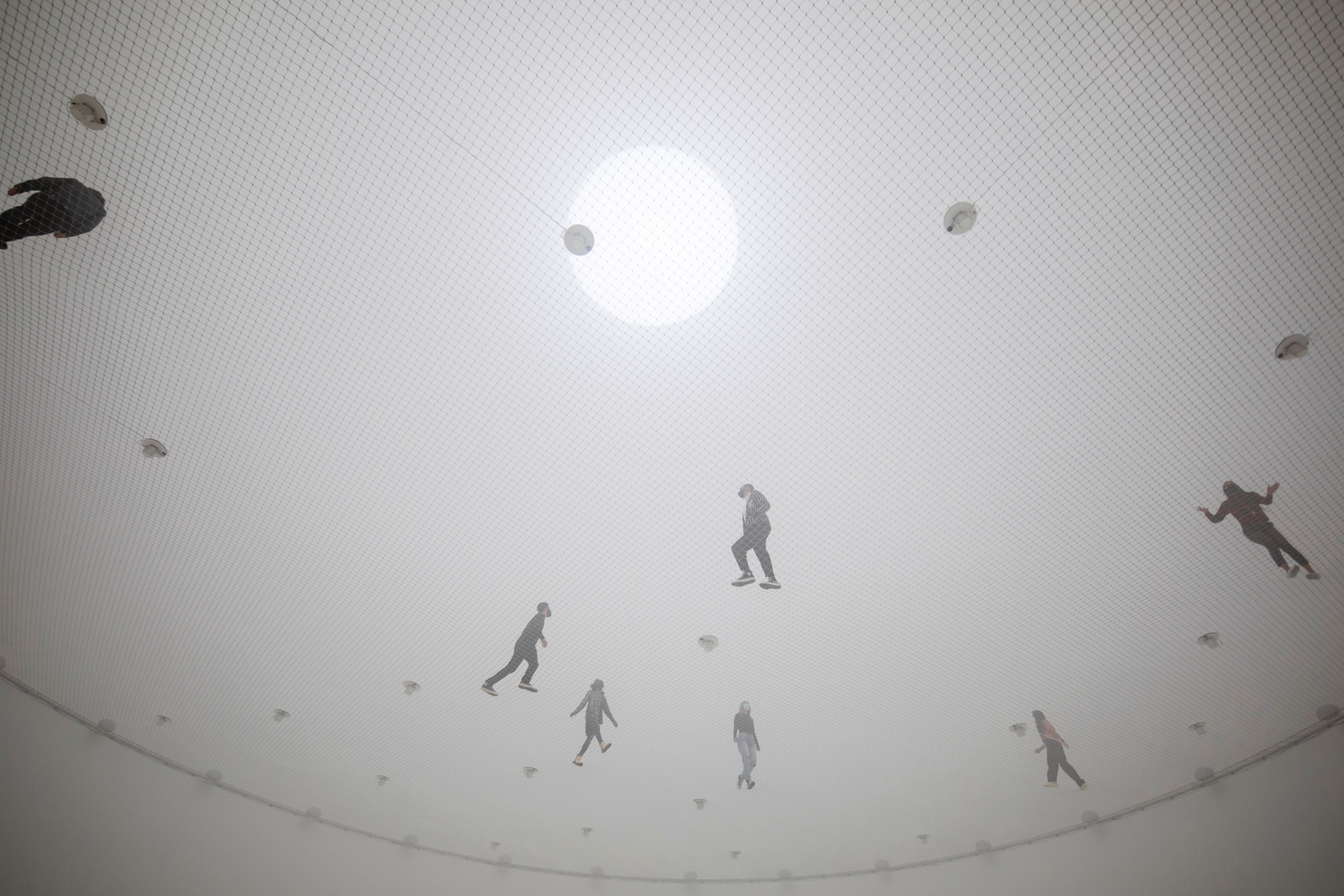 People walking on a net suspended 40 feet in the air. They are seen from below and are spaced out across the net, within a large white spherical space.
