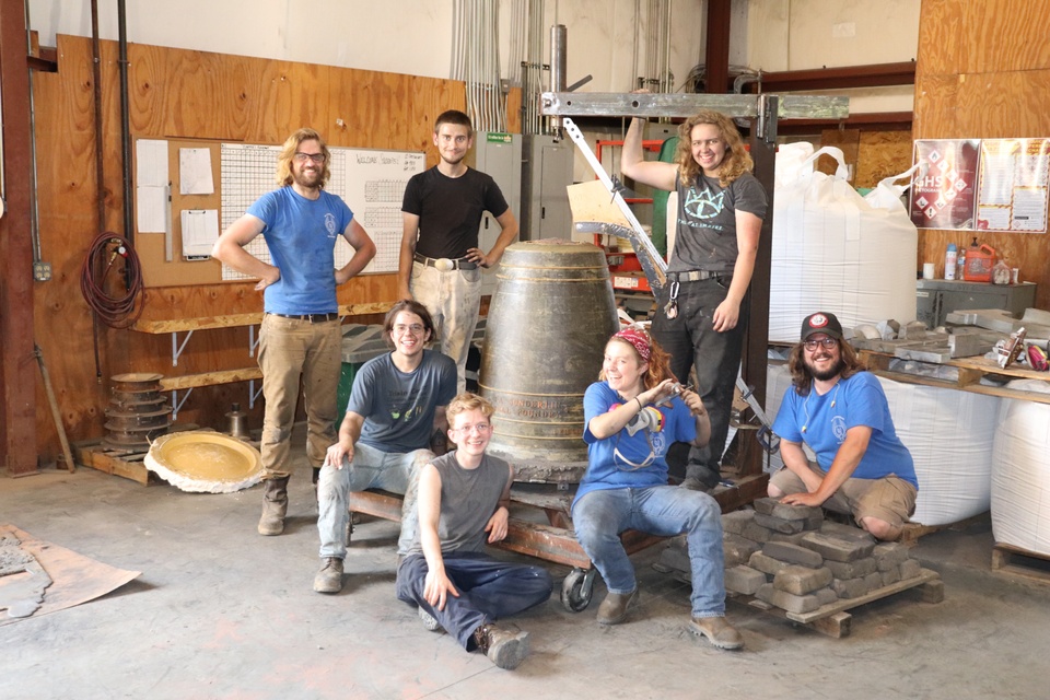 Group of seven people gathered around a bronze-cast bell at a foundry.