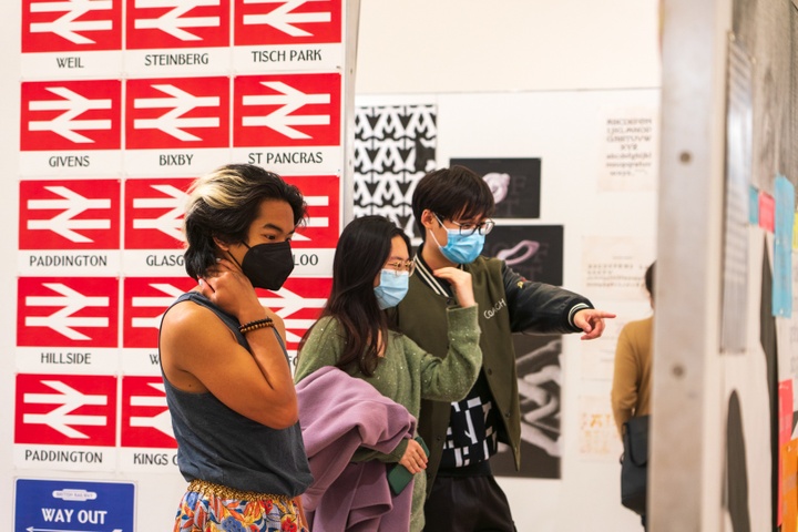 Students discuss works in a typographic gallery display. Behind them is a wall of type samples done in the style of train station signs.