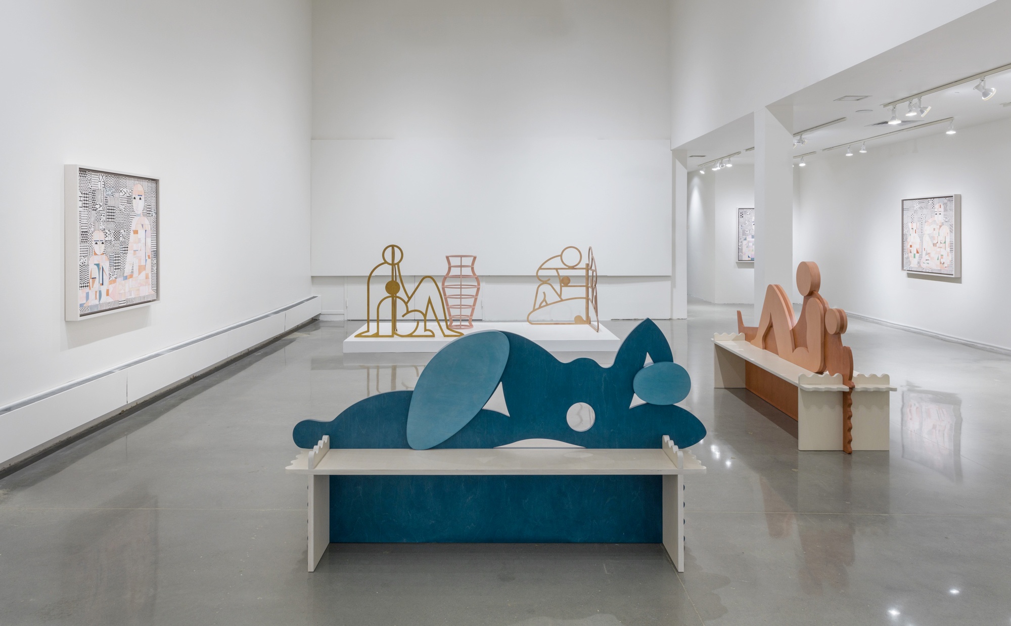 A sculpture of a large, simplistic, blue female figure reclines on a bench in front of another sculpture with multiple abstract figures sitting around a vase.