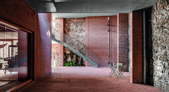 View of a covered, red-brick court space with a mix of brick and stone walls, and a metal staircase near an opening with light coming in.