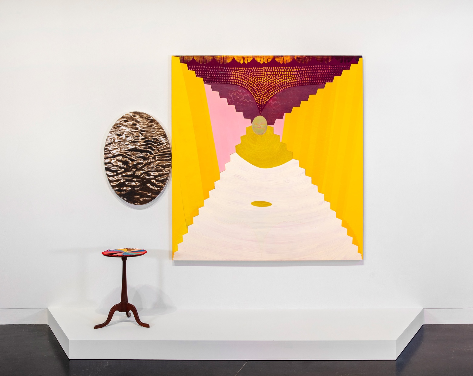 On a white wall, an oval artwork hangs next to a large, warm-colored painting with a small wooden end table beneath them.