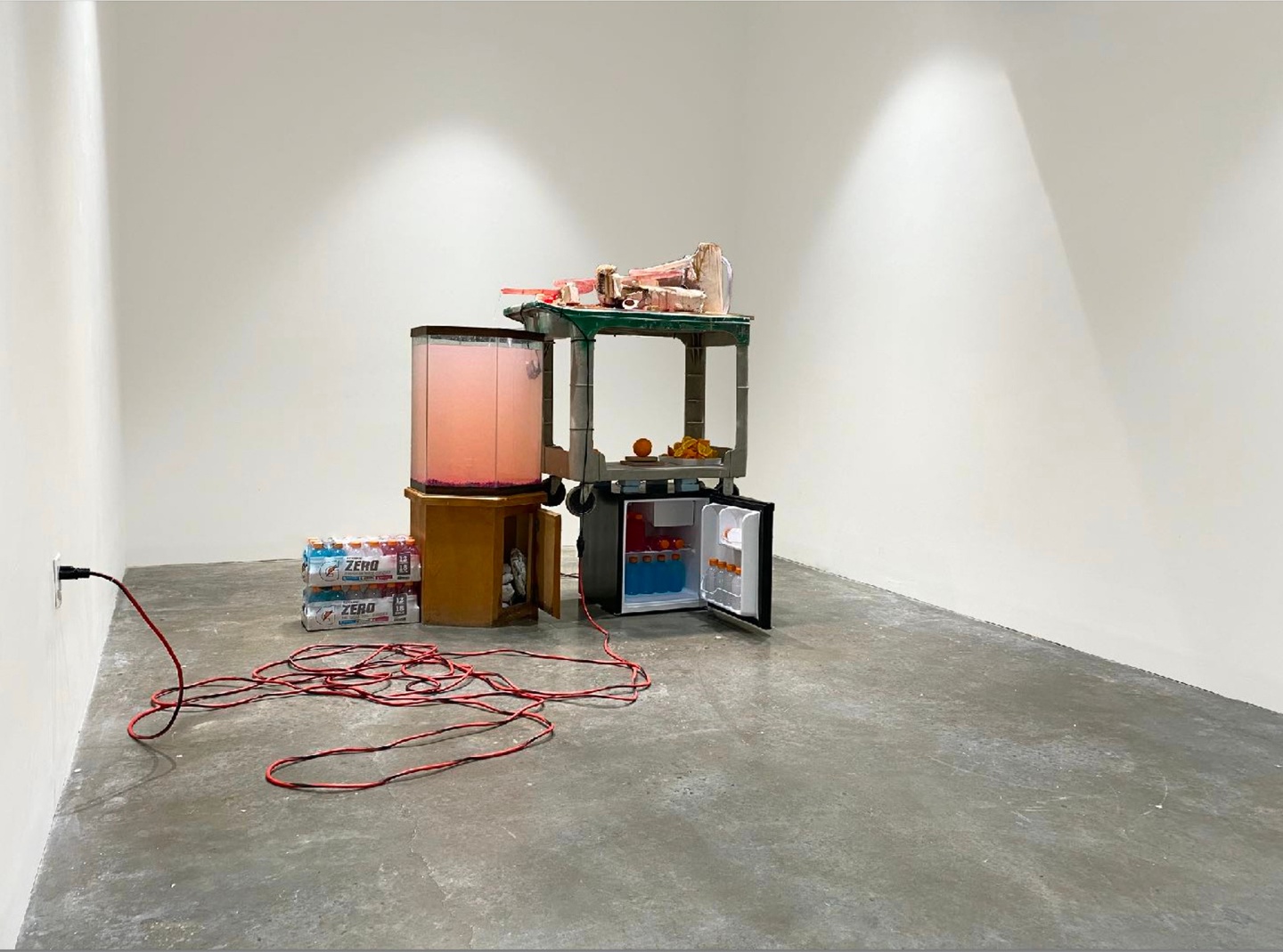 Mixed media sculpture featuring a mini-fridge plugged into a wall, filled with bottles of Gatorade; a cart is stacked on top, with oranges and clay on top. A wooden cabinet, door slightly ajar, with a clear cooler of red liquid are to the left, along with two small containers of Gatorade bottles.